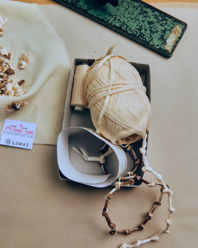 Overhead shot of a crafting box with woven raffia, spool of thread, fabric swatch with embroidery, and a small tagged label