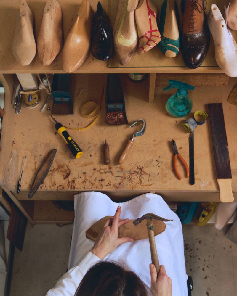 High-angle view of a shoemaker crafting footwear at a cluttered workbench with tools and wooden lasts for T Magazine China.