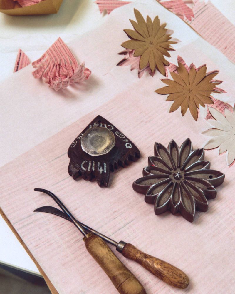 Overhead view of craft tools, carved wooden printing blocks, and cut paper flower shapes on pink fabric