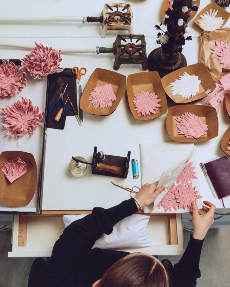 Overhead view of a workbench with paper craft materials, pink flower cutouts, tools, and hands working on paper art.