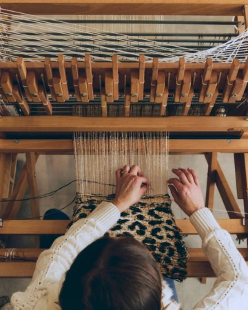High-angle shot of a person weaving on a wooden loom, working threads with hands while wearing a cream-colored knit sweater.