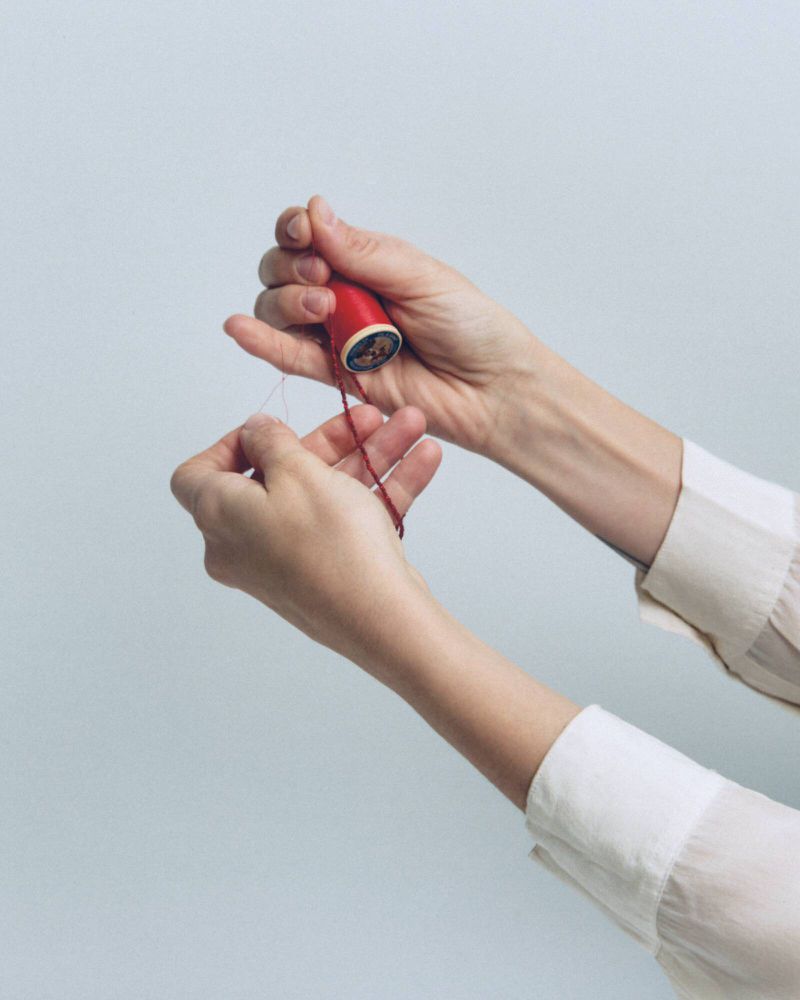 Close-up of hands holding a small red spool of thread with a trailing red string against a plain grey seamless background.