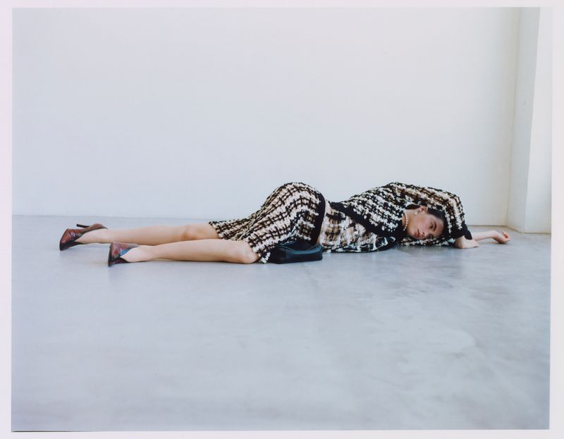 Model wearing patterned tweed outfit lying horizontally on a light grey floor against a plain wall