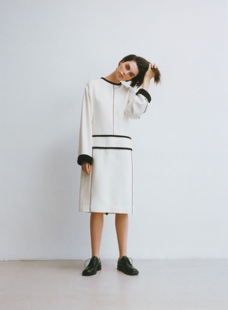 Model in a cream midi dress with black piping and dark shoes posing against a plain white background
