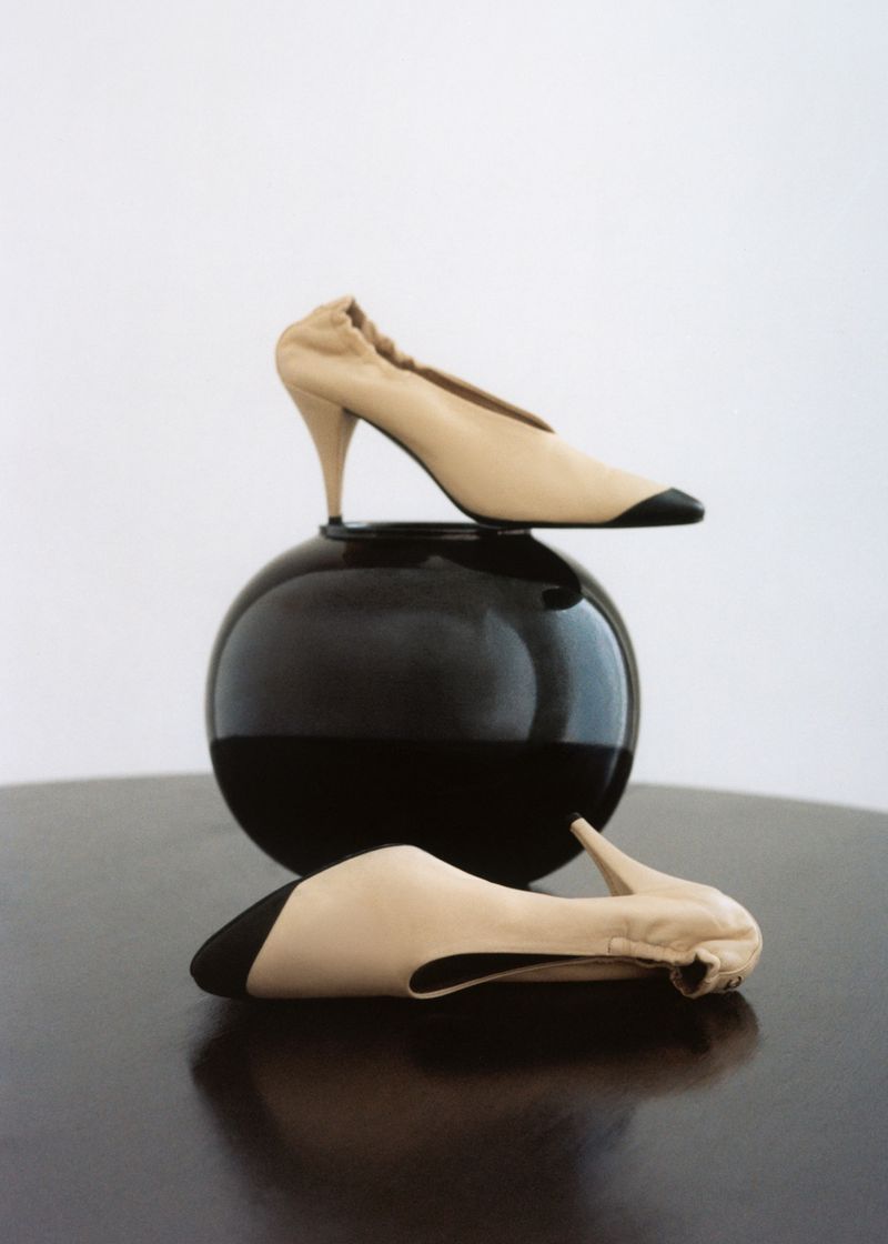 Beige high heel shoes with black toes, one resting on a dark reflective sphere and another on a table surface.