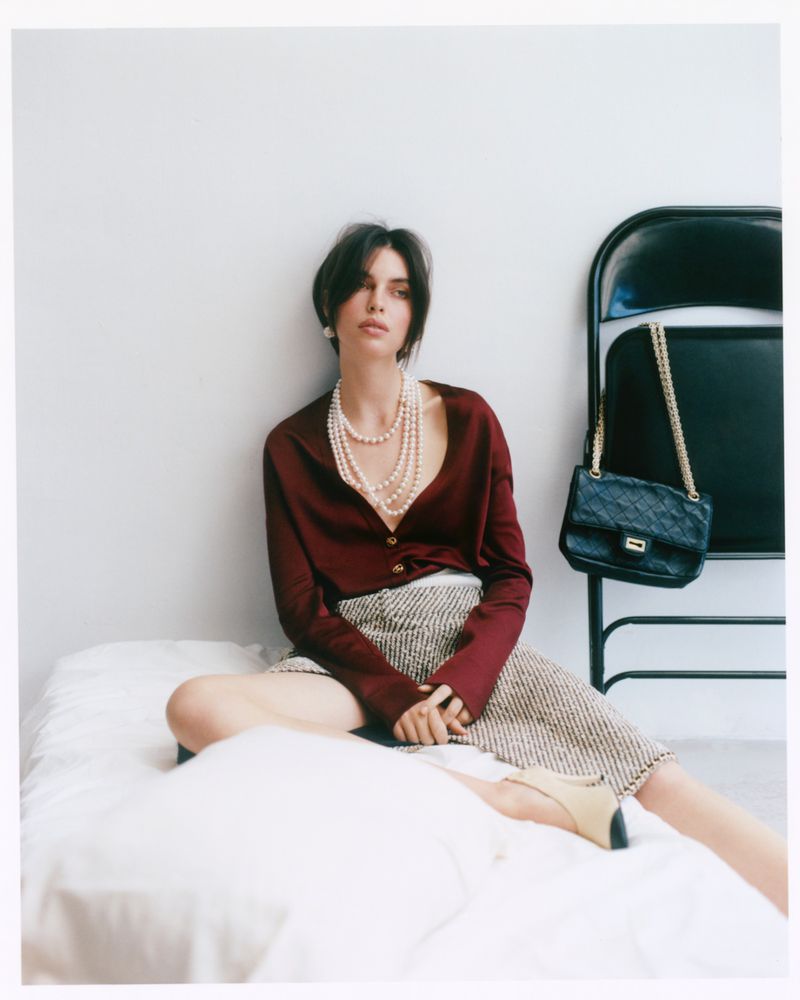 Model seated on a white bed wearing a burgundy blouse and patterned skirt with multi-strand pearl necklace, beside a black folding chair