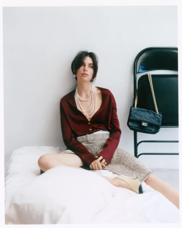 Model seated on a white bed wearing a burgundy blouse and patterned skirt with multi-strand pearl necklace, beside a black folding chair