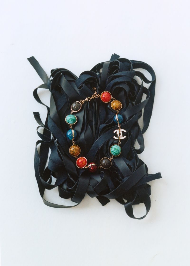 Colorful beaded necklace with a Chanel logo charm resting on a pile of dark navy ribbons against a white background.