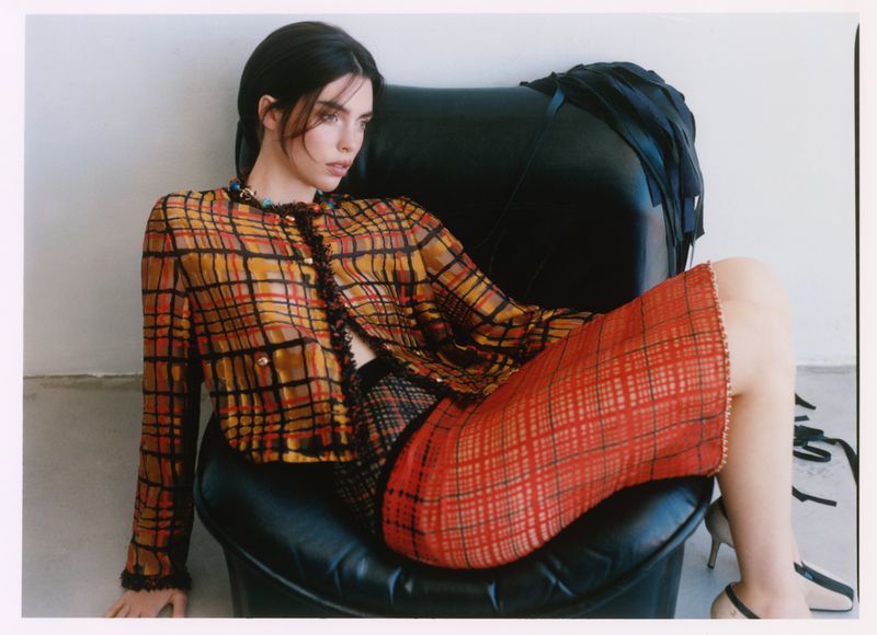Model wearing a plaid jacket and red patterned skirt seated on a black leather chair against a white wall.