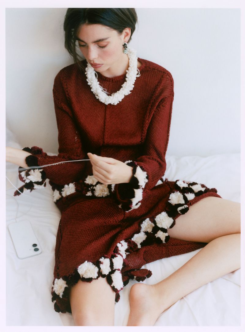 A young adult woman with dark hair wears a maroon textured dress and white flower necklace while seated on a white bed indoors.