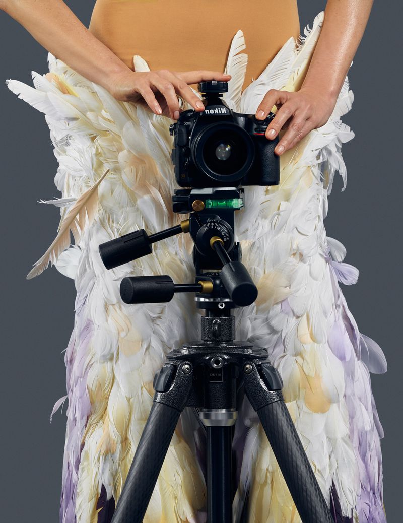Model wearing feathered skirt holding a professional DSLR camera mounted on a tripod against a grey studio backdrop.