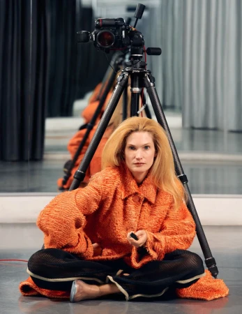 Model seated cross-legged in an orange textured jacket before a camera tripod with studio reflections