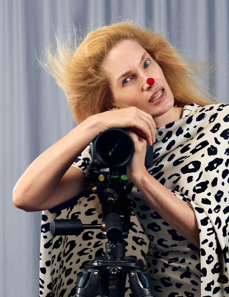 Model with red dot on nose wearing animal print garment leaning on professional camera tripod against grey background