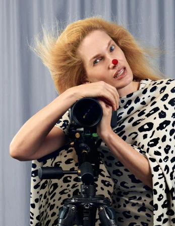 Model with red dot on nose wearing animal print garment leaning on professional camera tripod against grey background