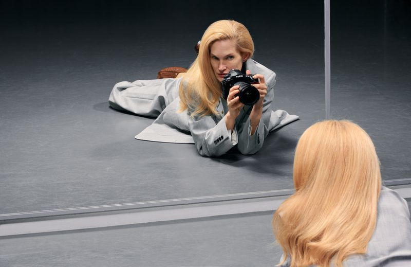 Blonde model in a grey coat lying on a grey floor holding a professional camera and looking into a mirror for Numero China