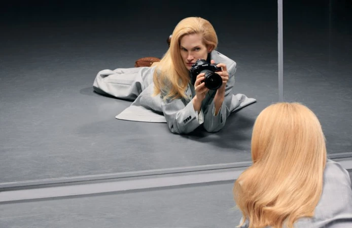 Blonde model in a grey coat lying on a grey floor holding a professional camera and looking into a mirror for Numero China
