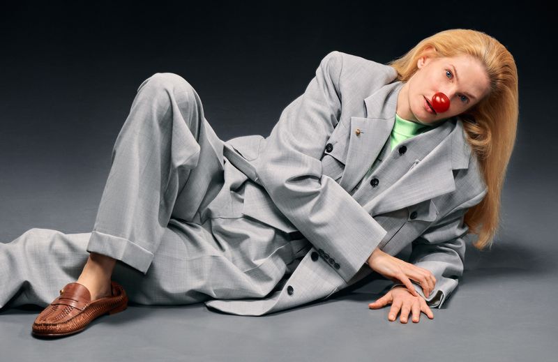 Model wearing an oversized grey suit and brown loafers reclining against a dark grey background with a red nose prop