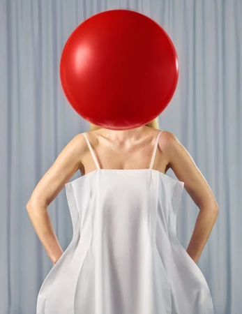 Model wearing a white structured dress with a large red balloon obscuring her face against grey curtains