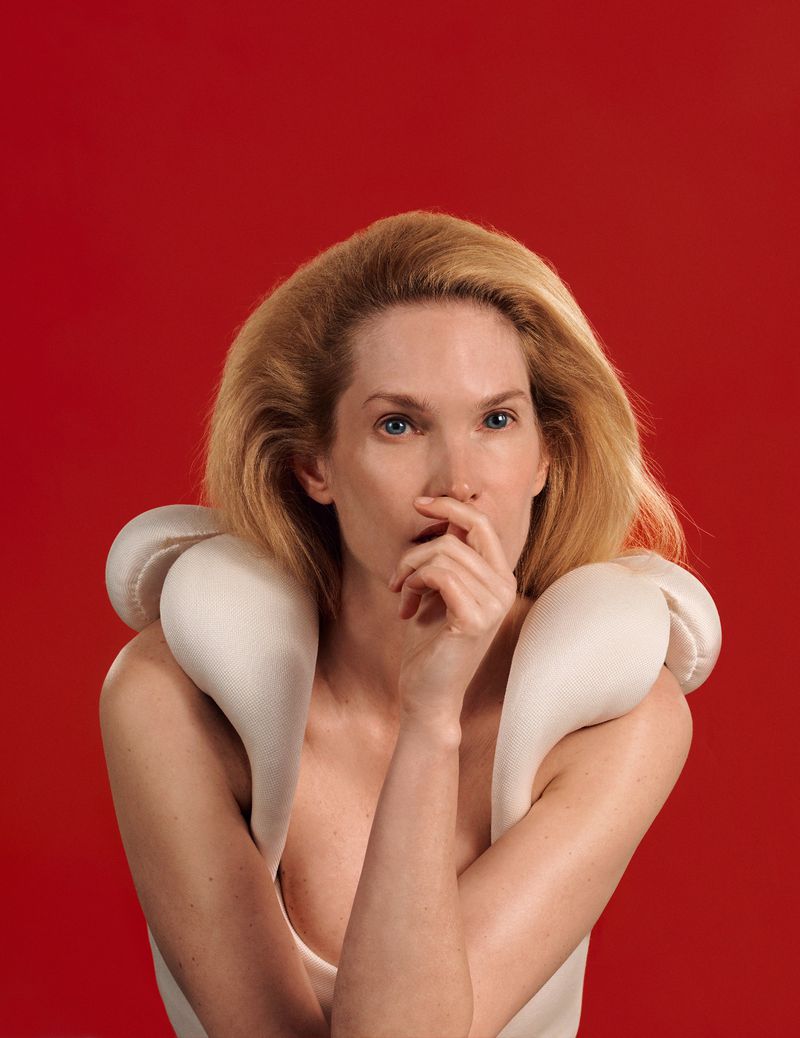 Portrait of a blonde woman with fair skin wearing an avant-garde padded white collar and dress against a red background.