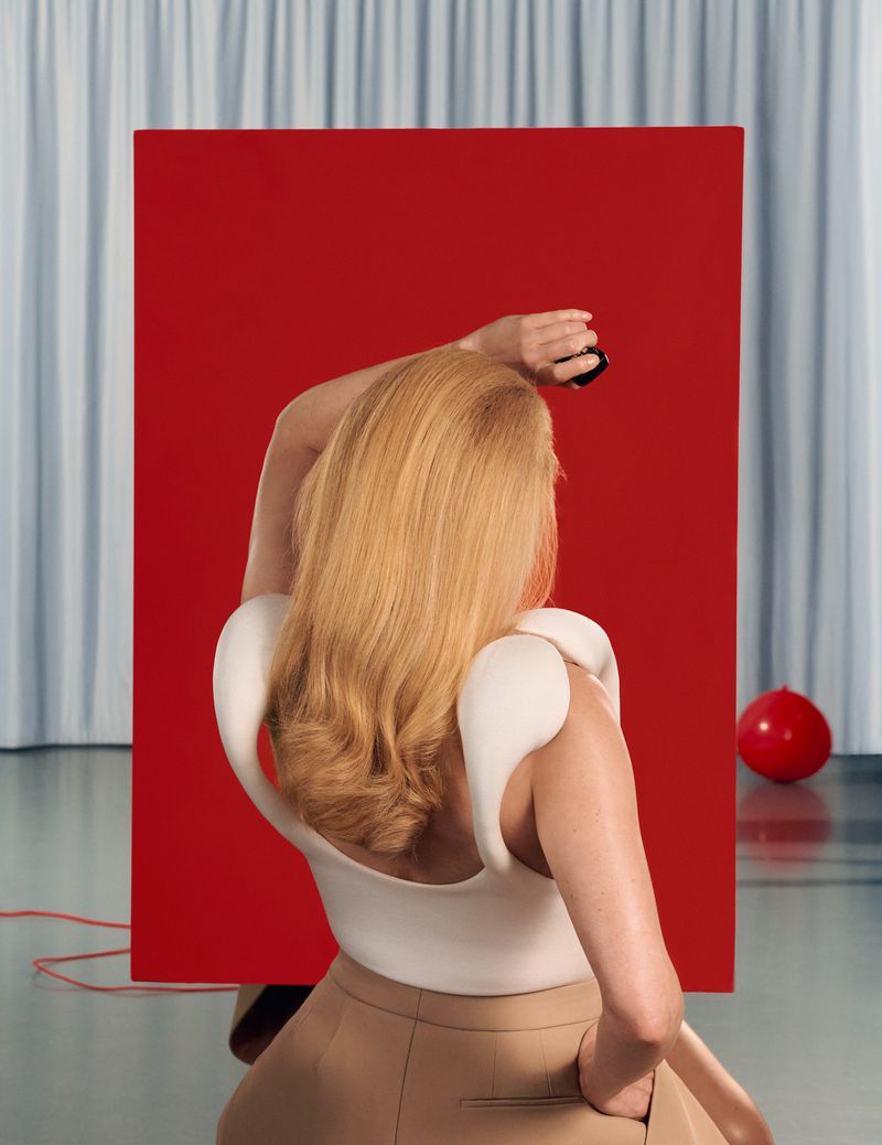 Blonde model seen from behind wearing white structured top and tan trousers against a red panel and grey curtain background.
