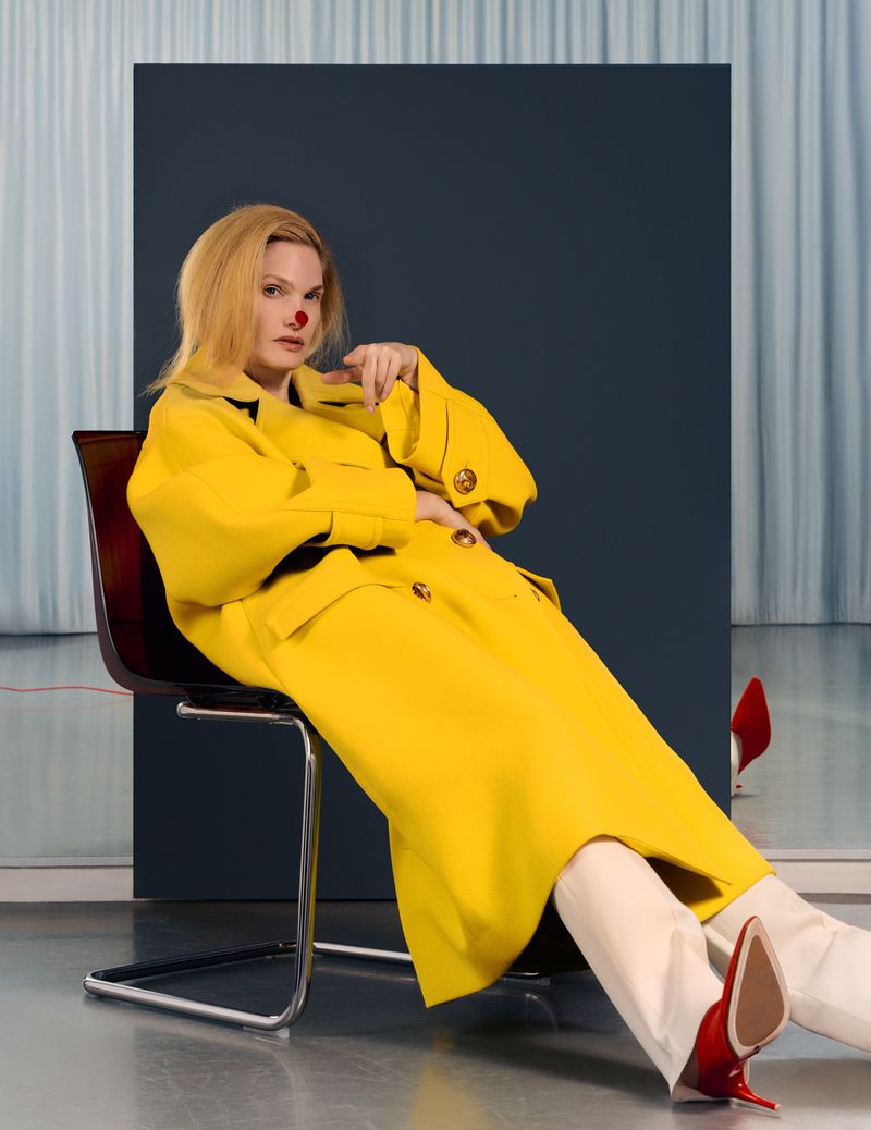 Model seated backwards on a chair wearing a bright yellow coat and red high heels against a backdrop for Numero China
