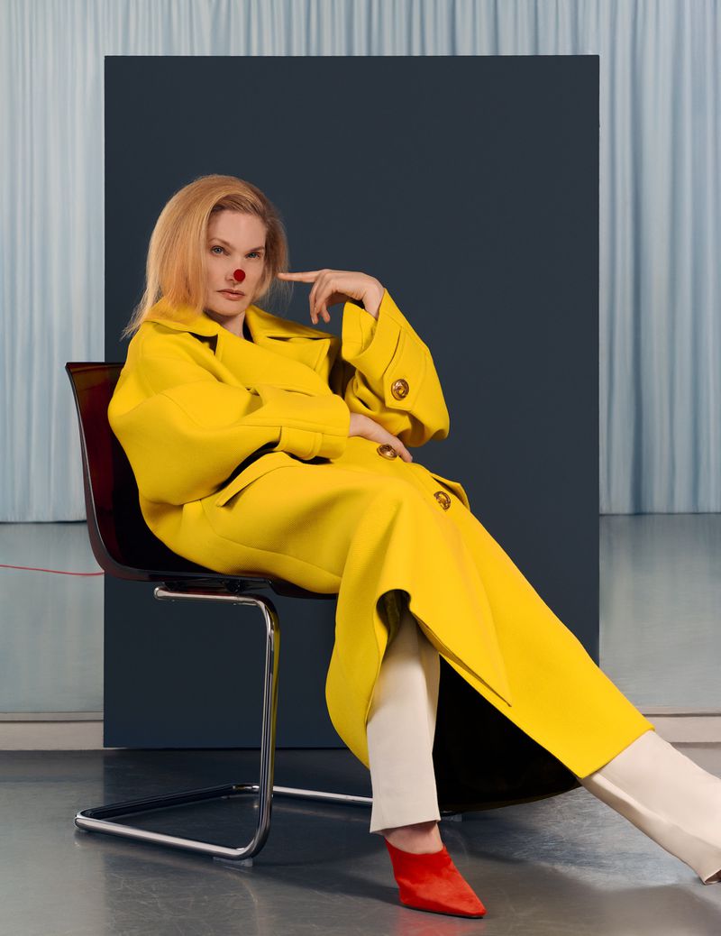 Model seated on a modern chair wearing a bright yellow coat and red shoes against a studio backdrop