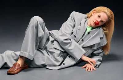 Model wearing an oversized grey suit and brown loafers reclining against a dark grey background with a red nose prop