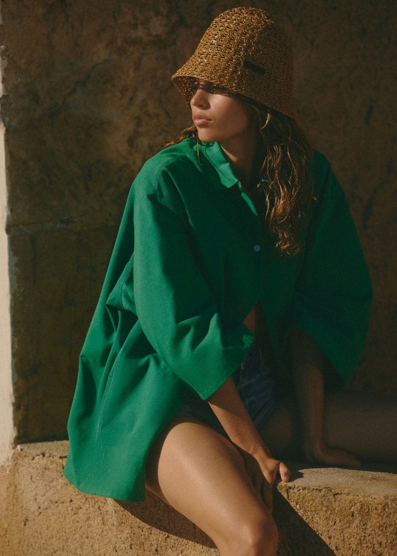 Model wearing a green button-down shirt and a straw bucket hat sitting on rough stone steps outdoors.