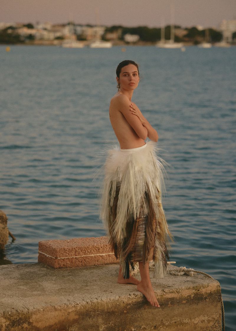 Model standing on a stone pier by the water wearing a textured feathered skirt for Elle France
