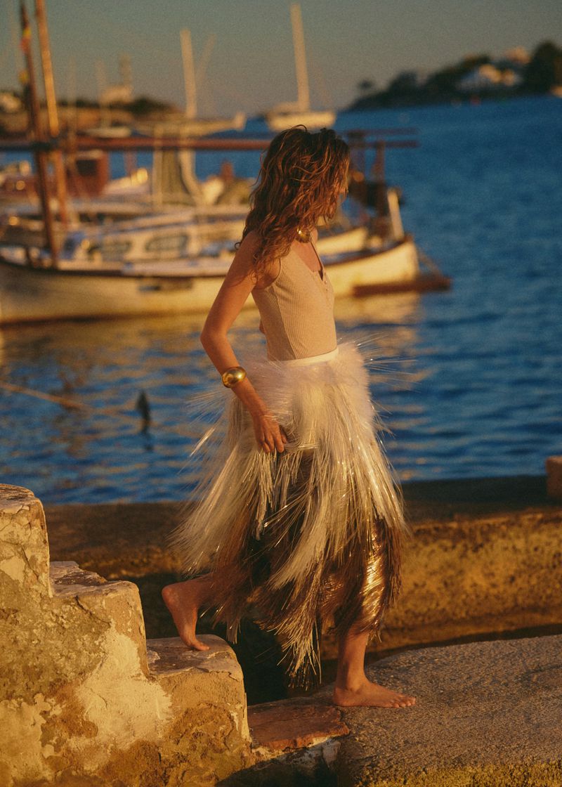 Model walking by a harbour at sunset wearing a beige top and textured fringe skirt