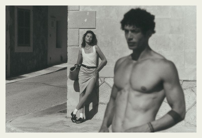 Black and white editorial fashion image featuring a standing woman leaning against a stone building wall and a shirtless man out of focus in the foreground