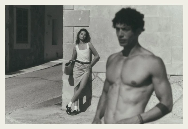 Black and white editorial fashion image featuring a standing woman leaning against a stone building wall and a shirtless man out of focus in the foreground