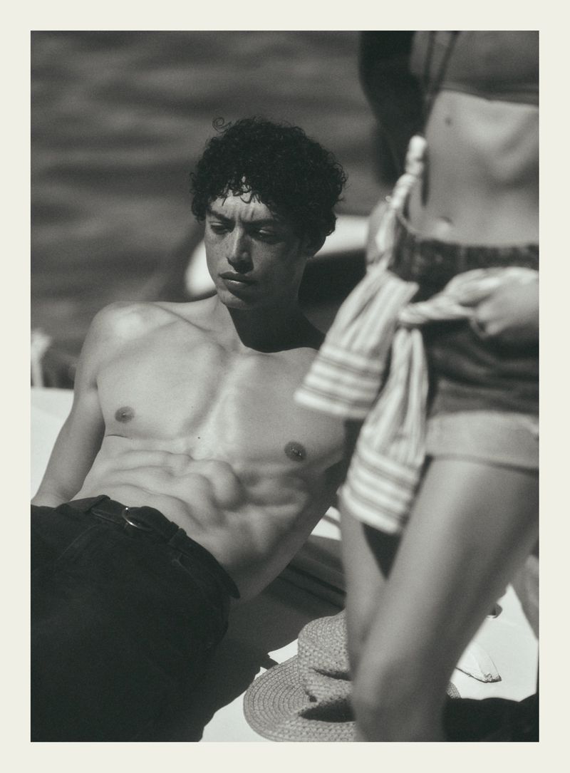 Black and white portrait of a shirtless man resting outdoors with a standing figure beside him on a sunny day