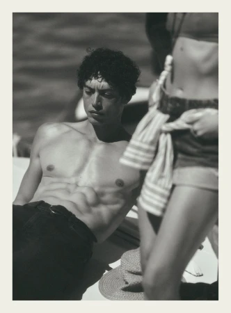 Black and white portrait of a shirtless man resting outdoors with a standing figure beside him on a sunny day