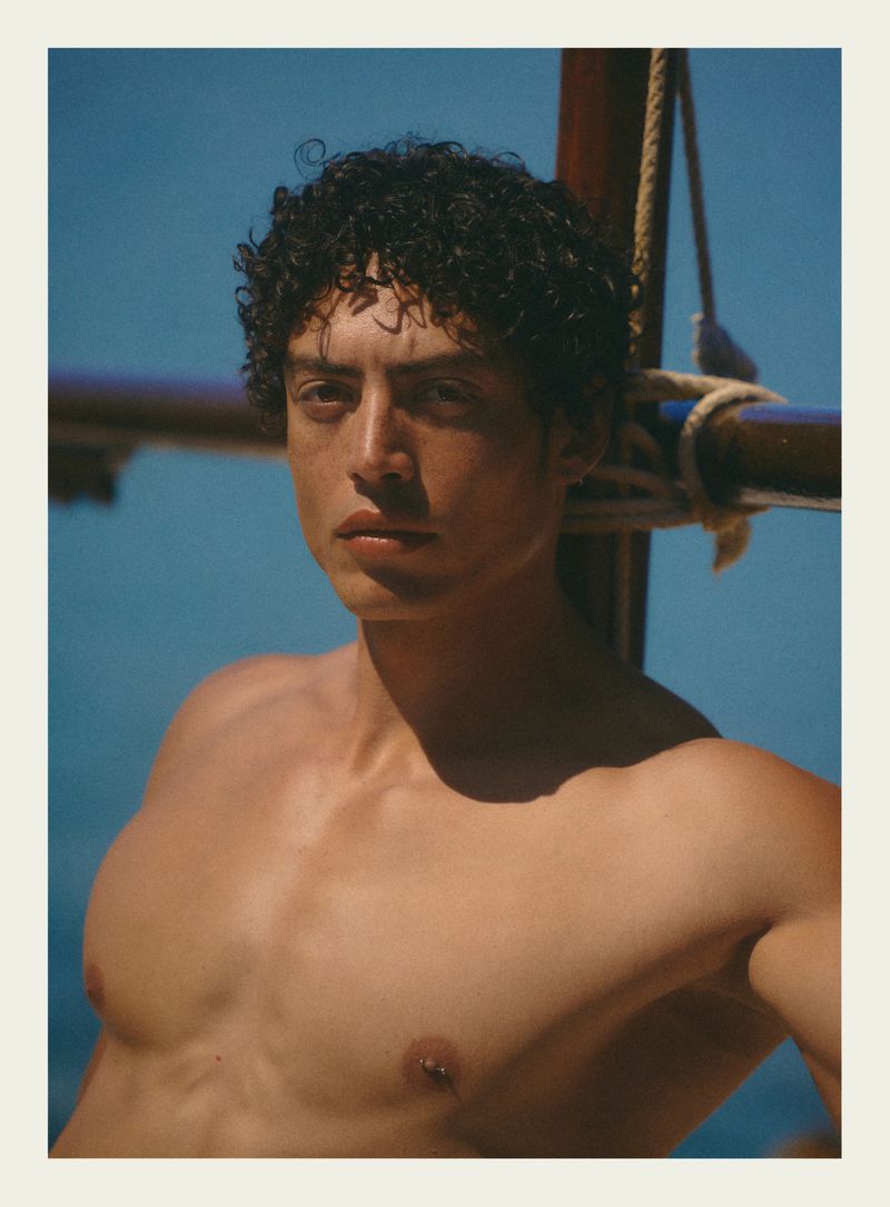 Close-up portrait of a shirtless male model with curly dark hair against a clear blue ocean background.