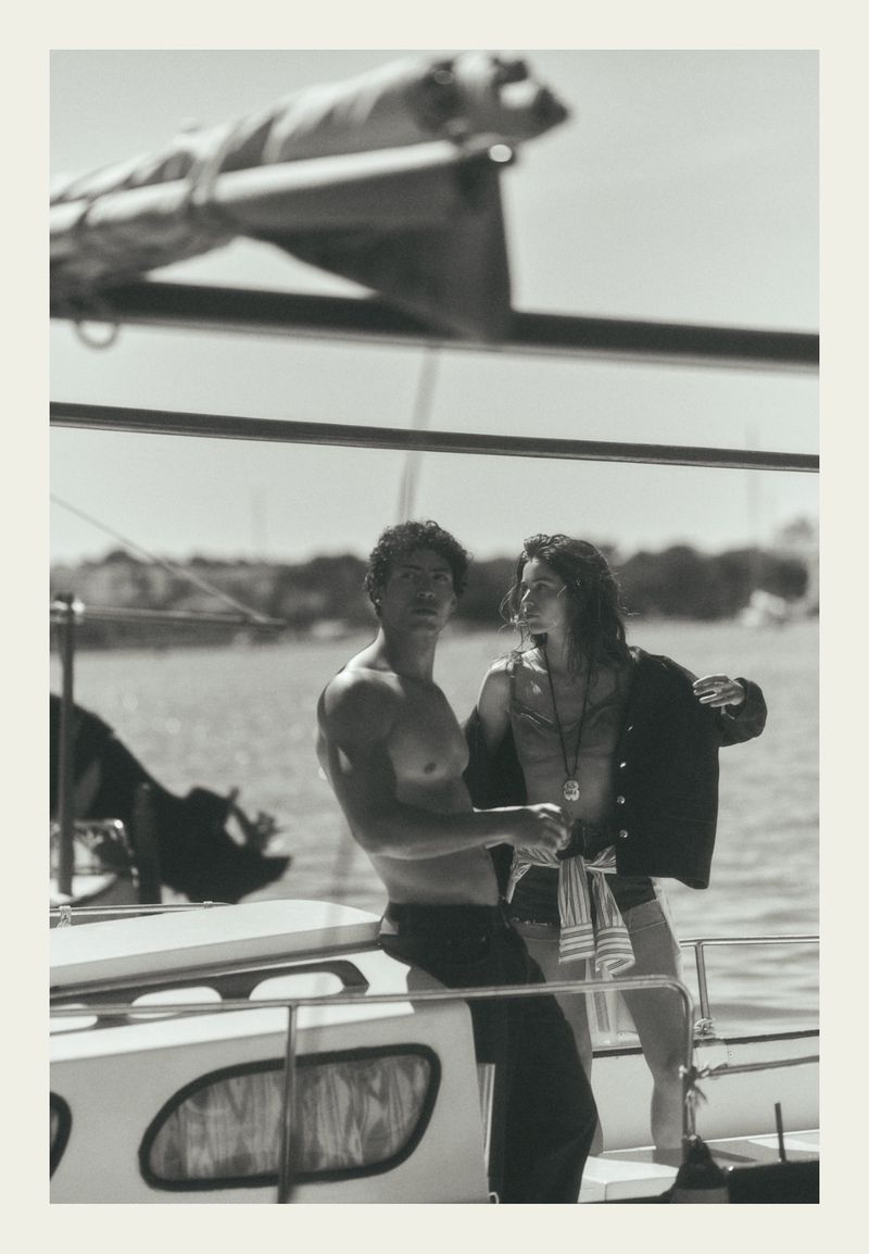 Black and white editorial photo of a man leaning on a sailboat and a woman standing beside him on deck