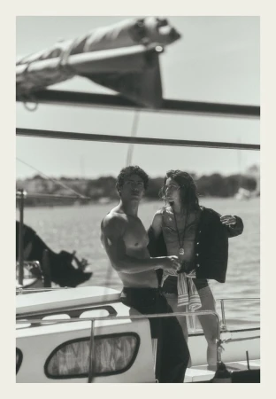 Black and white editorial photo of a man leaning on a sailboat and a woman standing beside him on deck