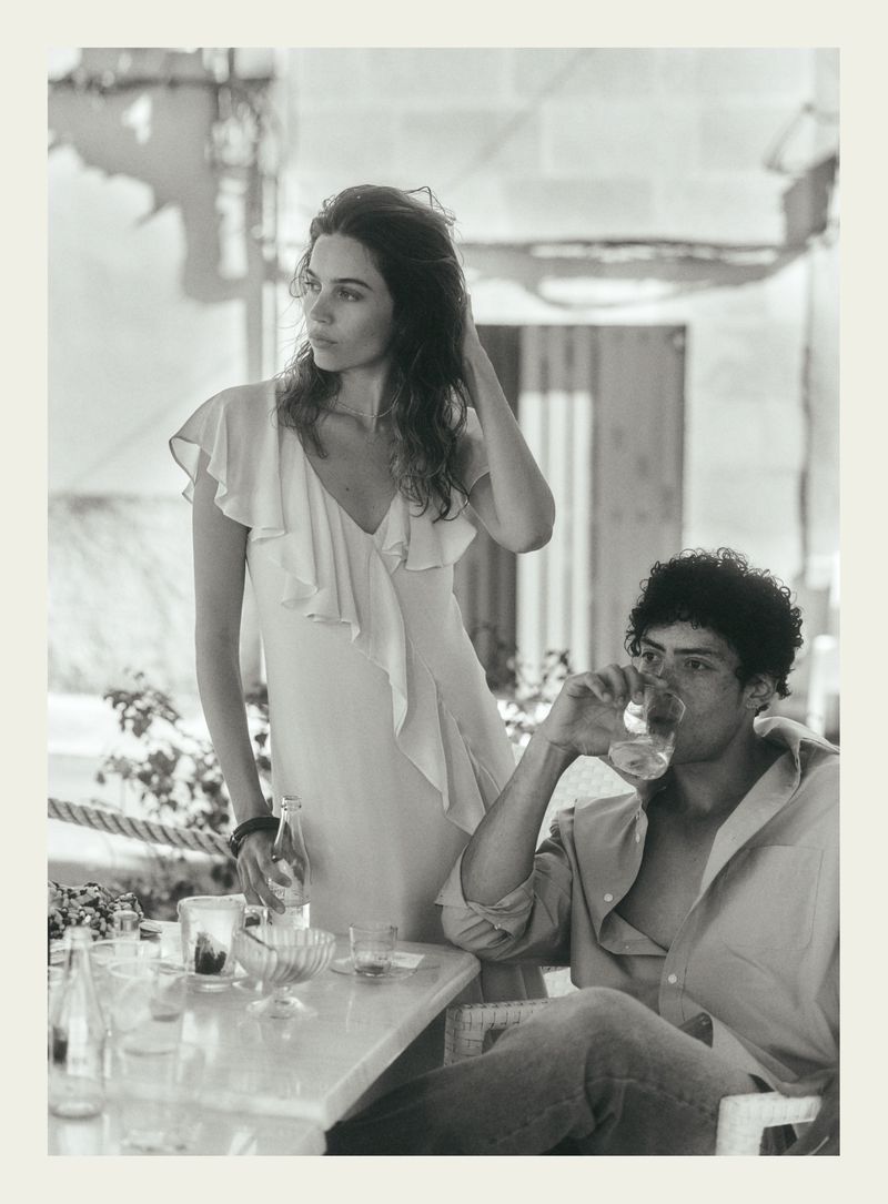 Black and white editorial image of a standing woman in a light dress and a seated man drinking from a glass at an outdoor table.