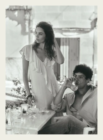 Black and white editorial image of a standing woman in a light dress and a seated man drinking from a glass at an outdoor table.