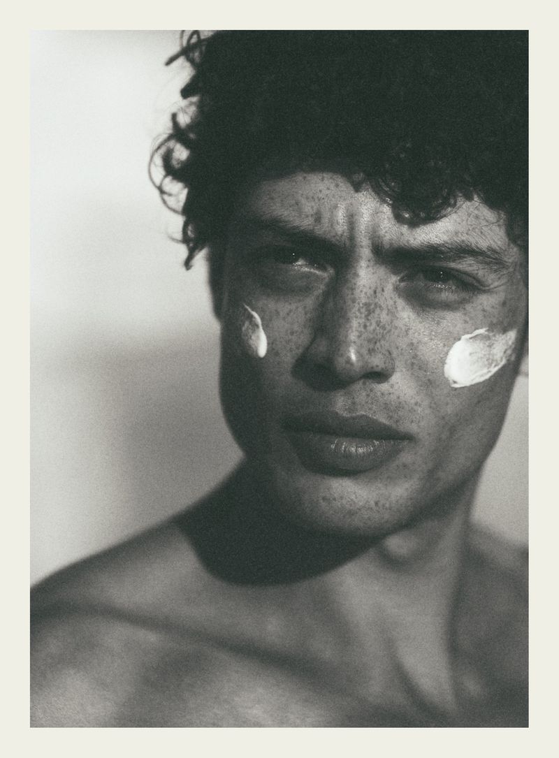 Close-up portrait of a male model with dark curly hair and freckles, wearing cream dabs on his cheeks, for Elle France.