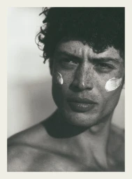 Close-up portrait of a male model with dark curly hair and freckles, wearing cream dabs on his cheeks, for Elle France.