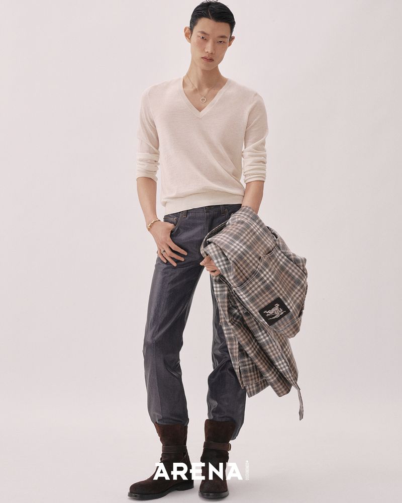Model wearing a cream v-neck sweater and dark trousers holding a plaid jacket against a seamless white background.
