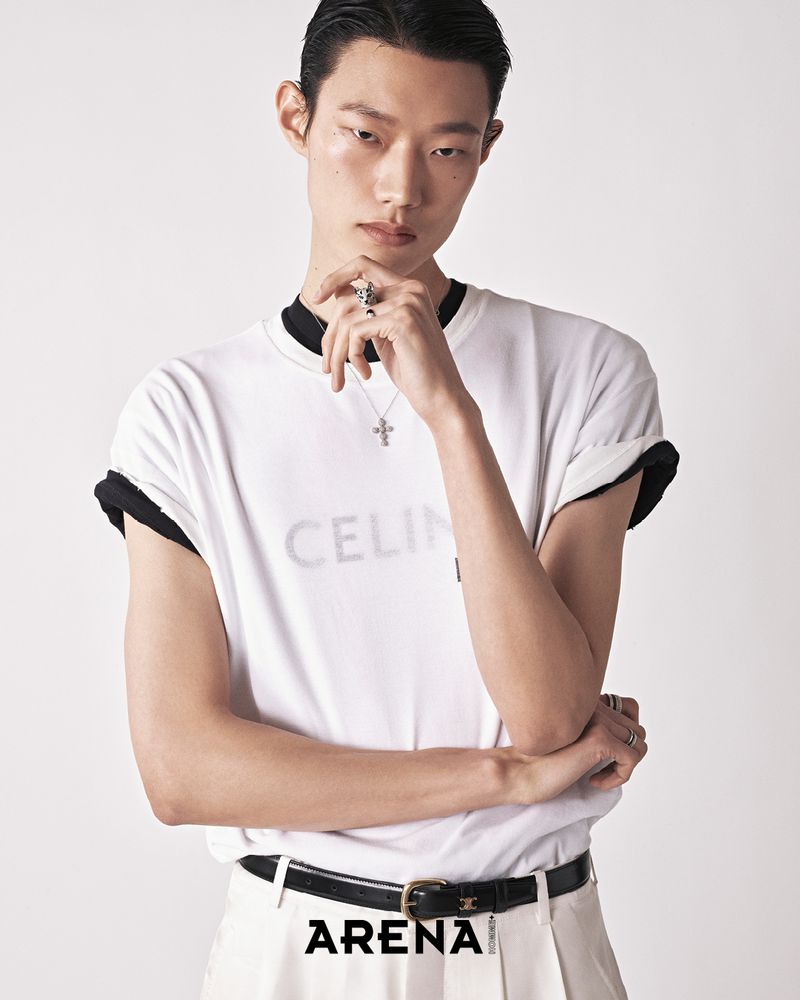 A stylish Asian male model poses in a white t-shirt and slacks, accessorized with jewelry, against a neutral backdrop in a studio setting.