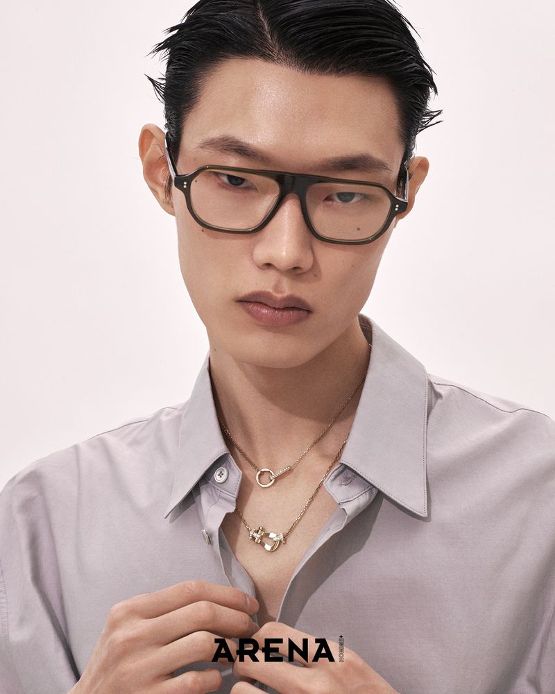 Asian man wearing tinted glasses and light grey collared shirt adjusts his buttons against a plain white background