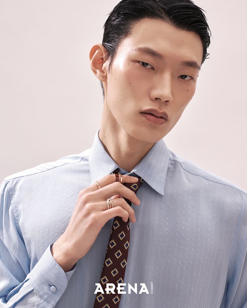 Close-up portrait of an East Asian male model in a light blue shirt adjusting a patterned dark tie against a plain background