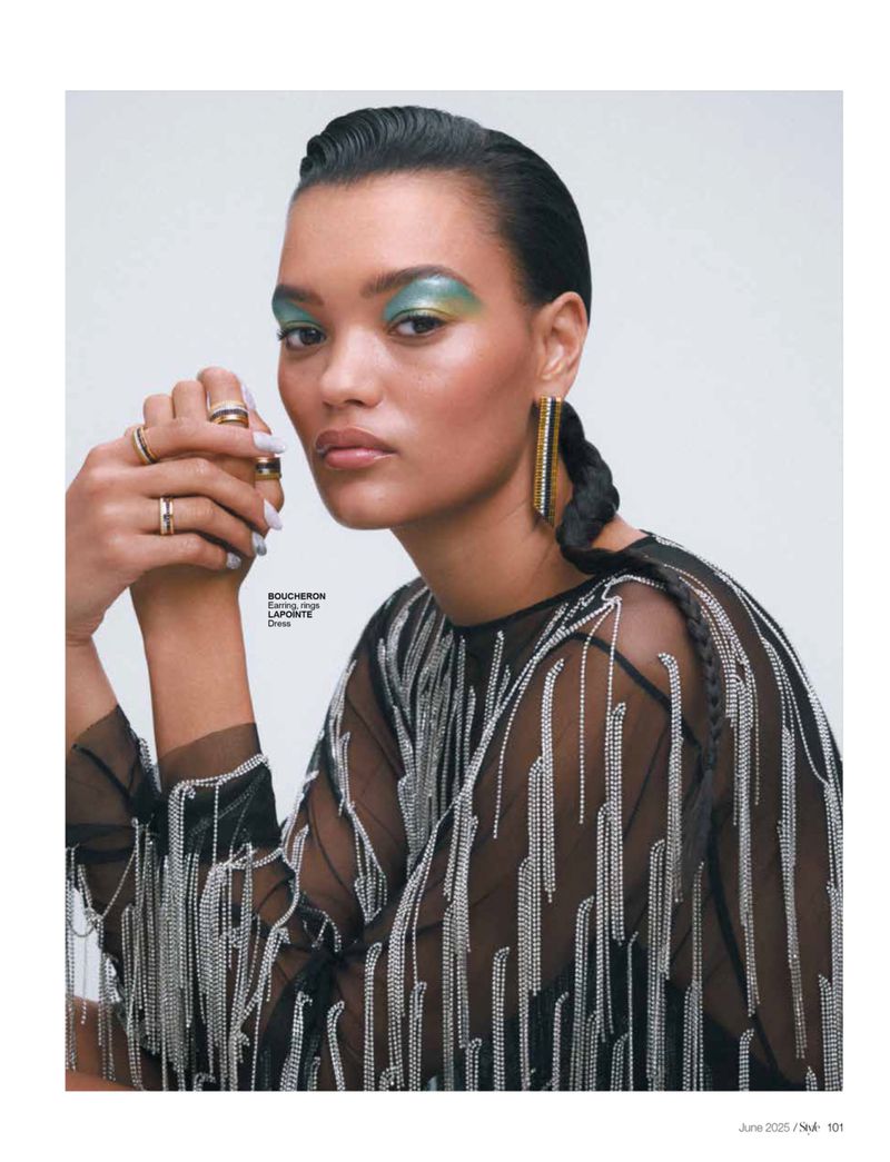 A striking portrait of a woman with bold eye makeup, statement earrings, and rings, styled in a sheer black top with silver fringe.