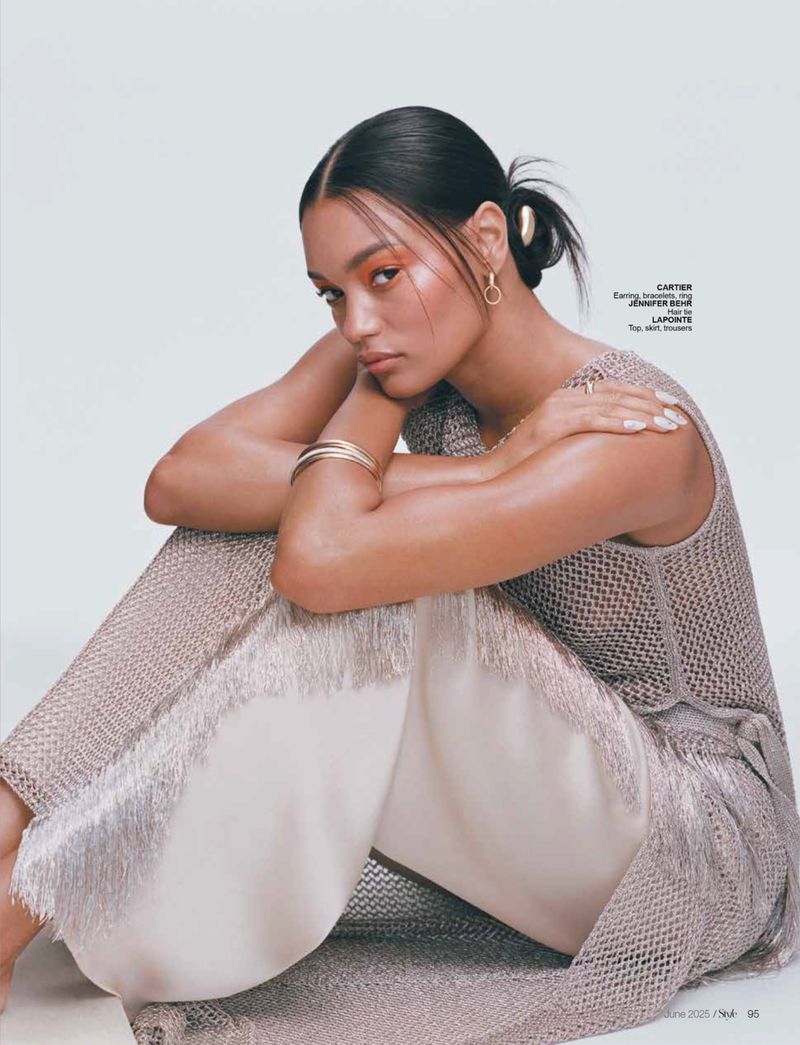 Woman in beige knit dress and ivory fringe pants seated against a white backdrop wearing gold earrings and a gold bracelet.