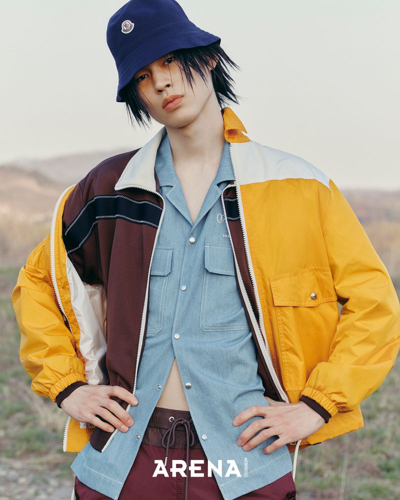 Model wearing a blue bucket hat, blue button-down shirt, and yellow and brown jacket outdoors.
