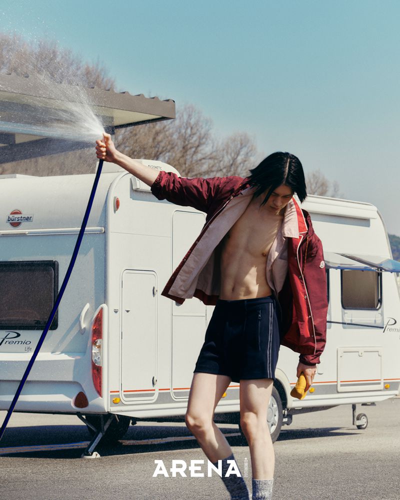 A man with dark hair holding a water hose outdoors in front of a white camper trailer for an Arena Homme + Korea editorial.
