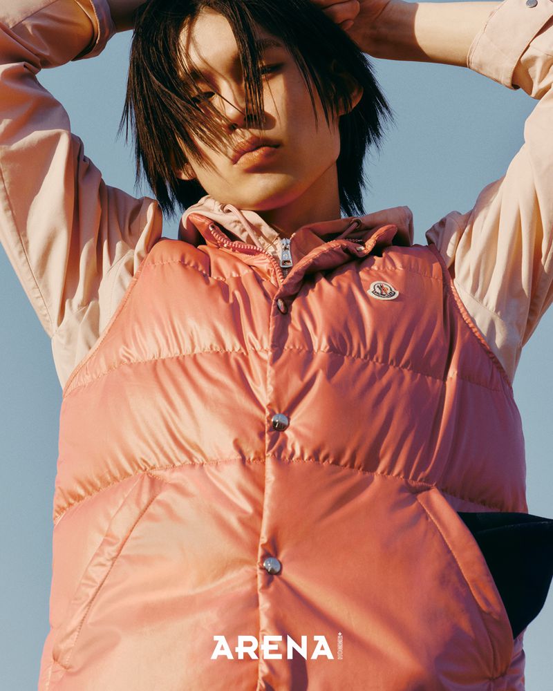 Model wearing a pink puffer vest and light pink hoodie with hands behind head against a clear sky background